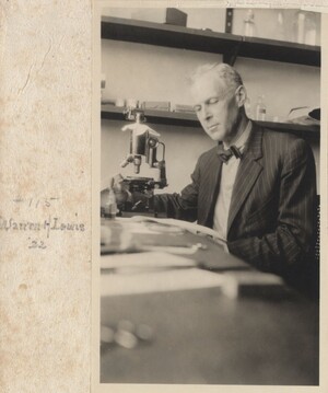 Lewis is seated at a table in front of a microscope, looking down at a paper. There are several pieces of paper and other items on the table, and there are shelves holding various boxes and glass containers on the far wall. He is wearing a pinstriped suit