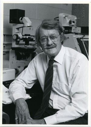 John P. Trinkaus wearing a long-sleeved white shirt, striped tie, and glasses, seated in front of a microscope